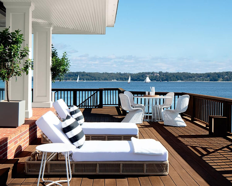 Spacious waterfront deck with white cushioned lounge chairs, black and white striped pillows, and a dining table with white wicker chairs, overlooking a calm blue lake with sailboats and a tree-lined shoreline in the distance.